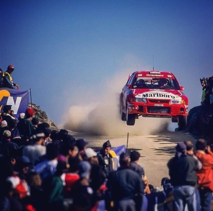 Red Mitsubishi Lancer Evolution rally car jumping on a gravel stage in front of spectators
